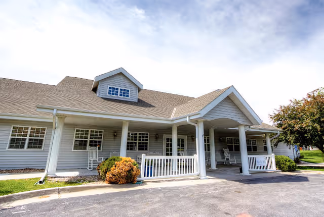 Exterior front view of a single-story senior living facility building with gray siding, white trim, a covered entrance supported by columns, rocking chairs on the porch, and some bushes and trees around the building.