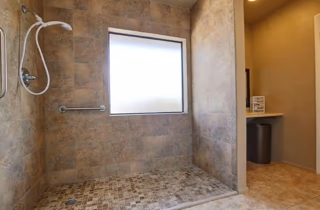 A walk-in shower with beige and brown tiled walls and floor, featuring a handheld showerhead and a horizontal grab bar mounted on the wall. There is a frosted window providing natural light, and an adjacent area with a countertop and trash bin visible.