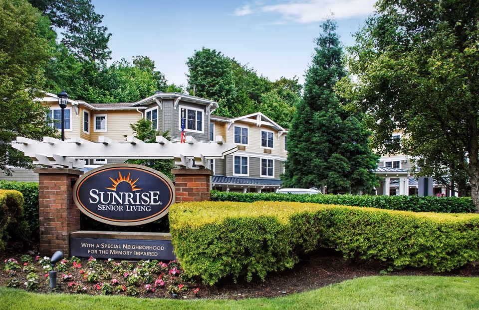 Exterior view of Sunrise Senior Living facility with a large sign in front surrounded by landscaped bushes and flowers, and a multi-story building with beige and gray siding in the background under a partly cloudy sky.