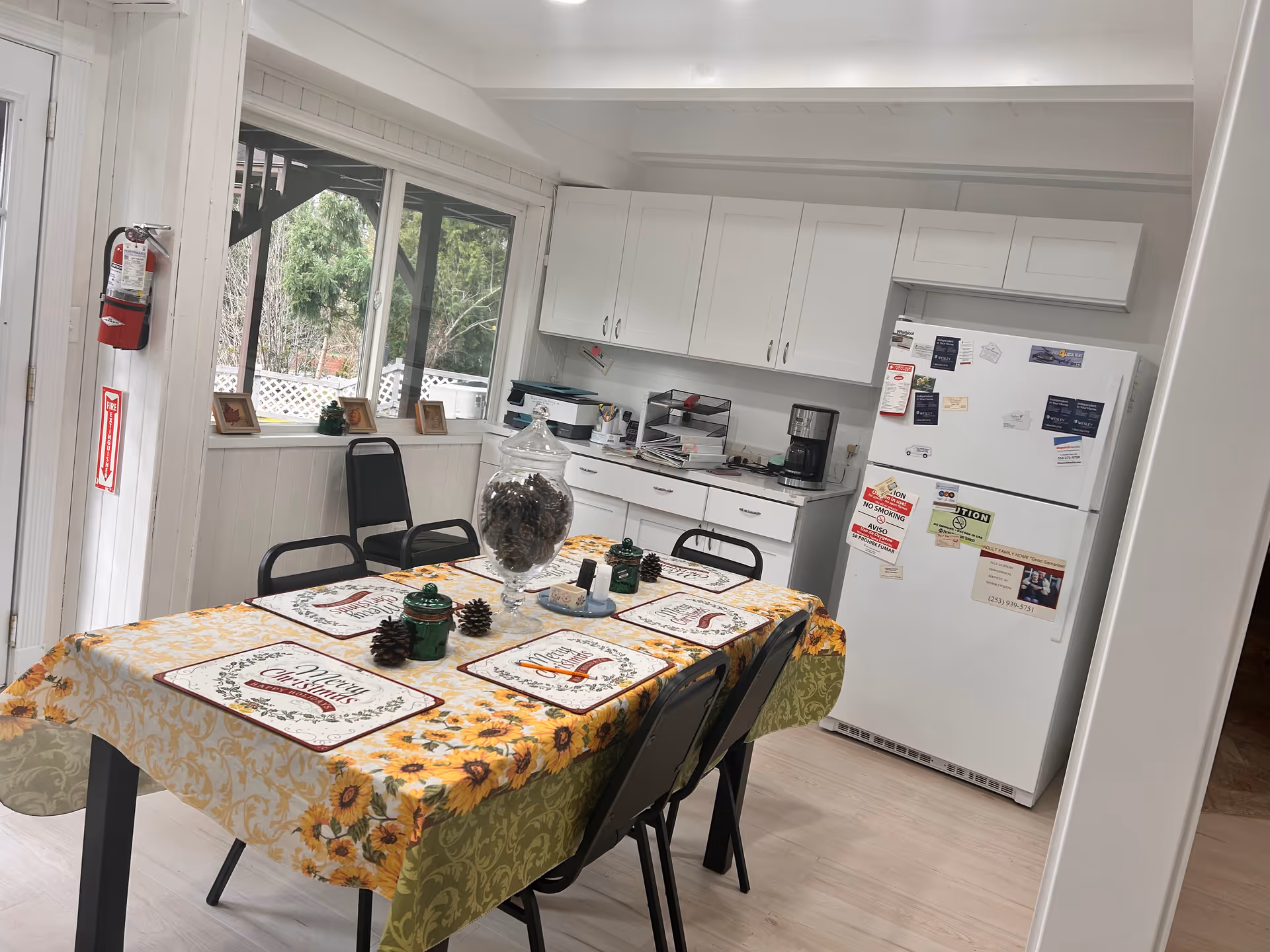 A bright kitchen area with white cabinets, a white refrigerator covered with various magnets and notices, a coffee maker on the counter, and a table covered with a sunflower-patterned tablecloth and Christmas-themed placemats. Four black chairs surround the table, and a large glass jar filled with pinecones is placed in the center. A fire extinguisher is mounted on the wall near a window that looks out to greenery outside.