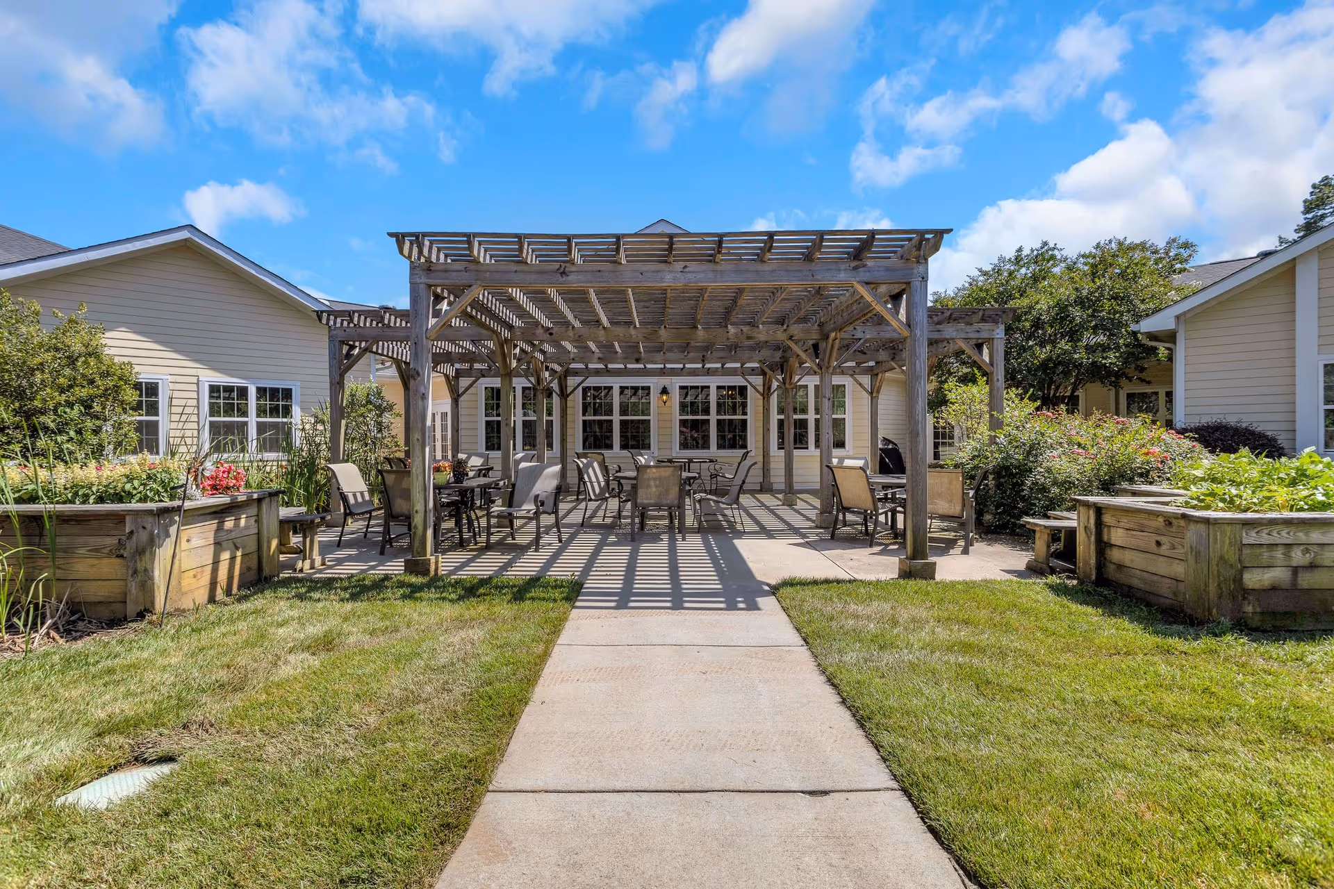 Outdoor patio area at Cadence at Clemmons featuring a wooden pergola with several tables and chairs underneath, surrounded by greenery and raised garden beds, with beige buildings in the background under a blue sky with some clouds.
