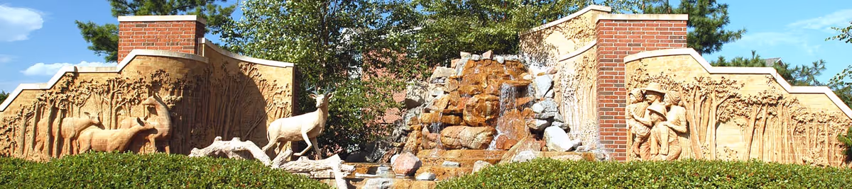 Landscaped entrance wall with carved wildlife and human reliefs flanking a central rock waterfall and brick pillars.