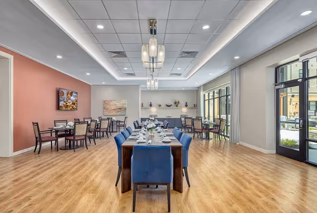 A spacious dining room with wooden flooring and modern lighting fixtures. The room features a long dining table with blue upholstered chairs set with white napkins and tableware. Additional smaller tables with chairs are arranged along the walls. Large windows and glass doors on the right side allow natural light to enter, and there is a view of an outdoor patio area.
