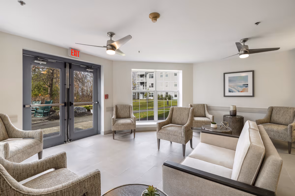 Bright, neutral-toned senior living lounge with sofas and armchairs arranged around coffee tables, a large window, and double glass exit doors.