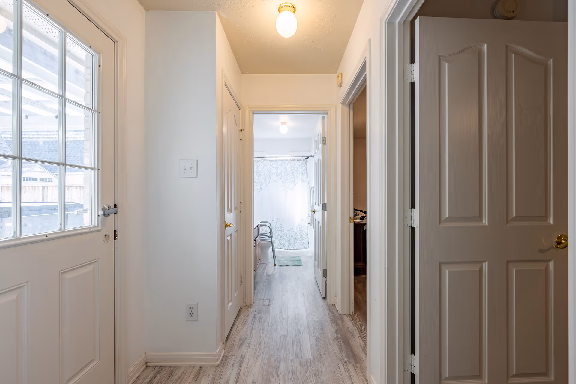 Hallway inside an assisted living residence with a glass-panel door on the left, light wood flooring, multiple open doors, and a bathroom with a shower curtain and walker at the far end.
