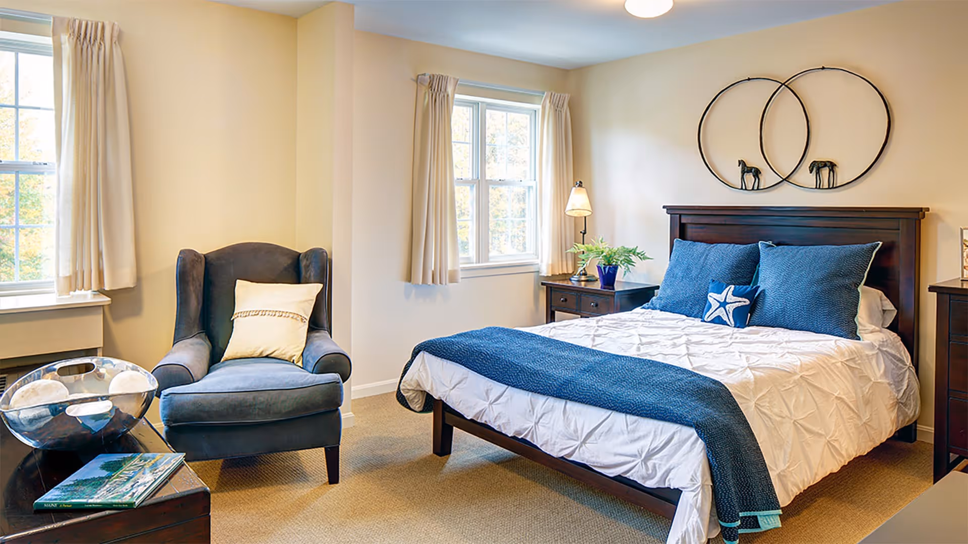 A cozy bedroom with a dark wooden bed frame, white bedding, and blue pillows and throw blanket. There is a blue armchair with a beige pillow next to a small table holding a decorative bowl and a book. Two windows with beige curtains let in natural light. On the wall above the bed are two circular wall decorations with small horse figurines inside.