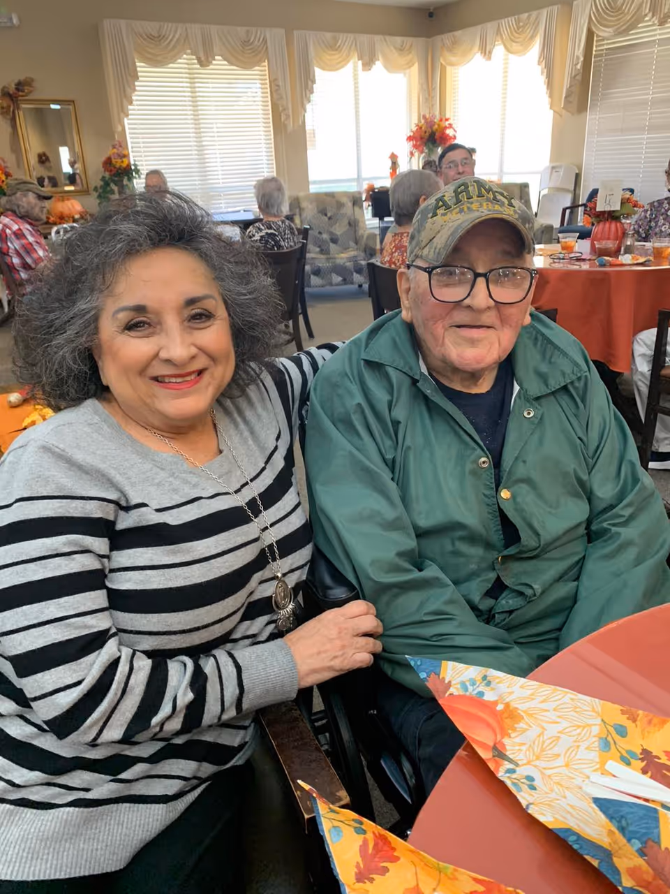 Two smiling older adults sit together at a decorated communal dining table in a bright activity room.