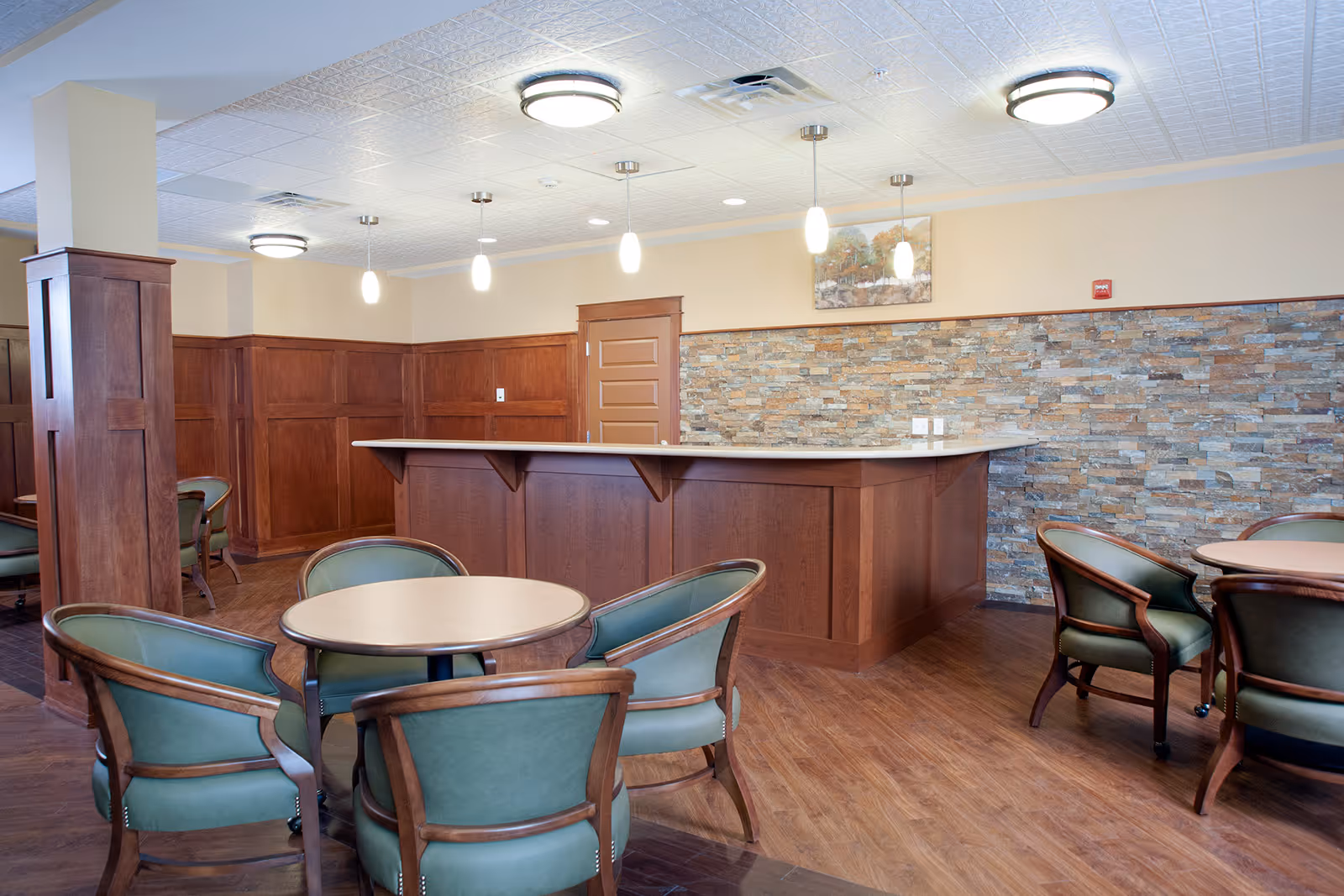 A cozy interior room with round tables and green cushioned chairs arranged around them. There is a wooden counter with a stone accent wall behind it, pendant lights hanging from the ceiling, and wood paneling on the walls. The floor is wooden, and the room has a warm, inviting atmosphere.