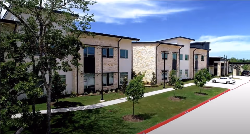 Exterior view of a modern two-story senior living facility building with stone and stucco facade, surrounded by green grass, small trees, and a sidewalk. Several parked cars are visible along the driveway under a clear blue sky.