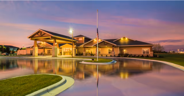 Exterior view of Oak Pointe of Monett senior living facility at dusk with a well-lit entrance, a flagpole in front, and a curved driveway reflecting the building and sky.