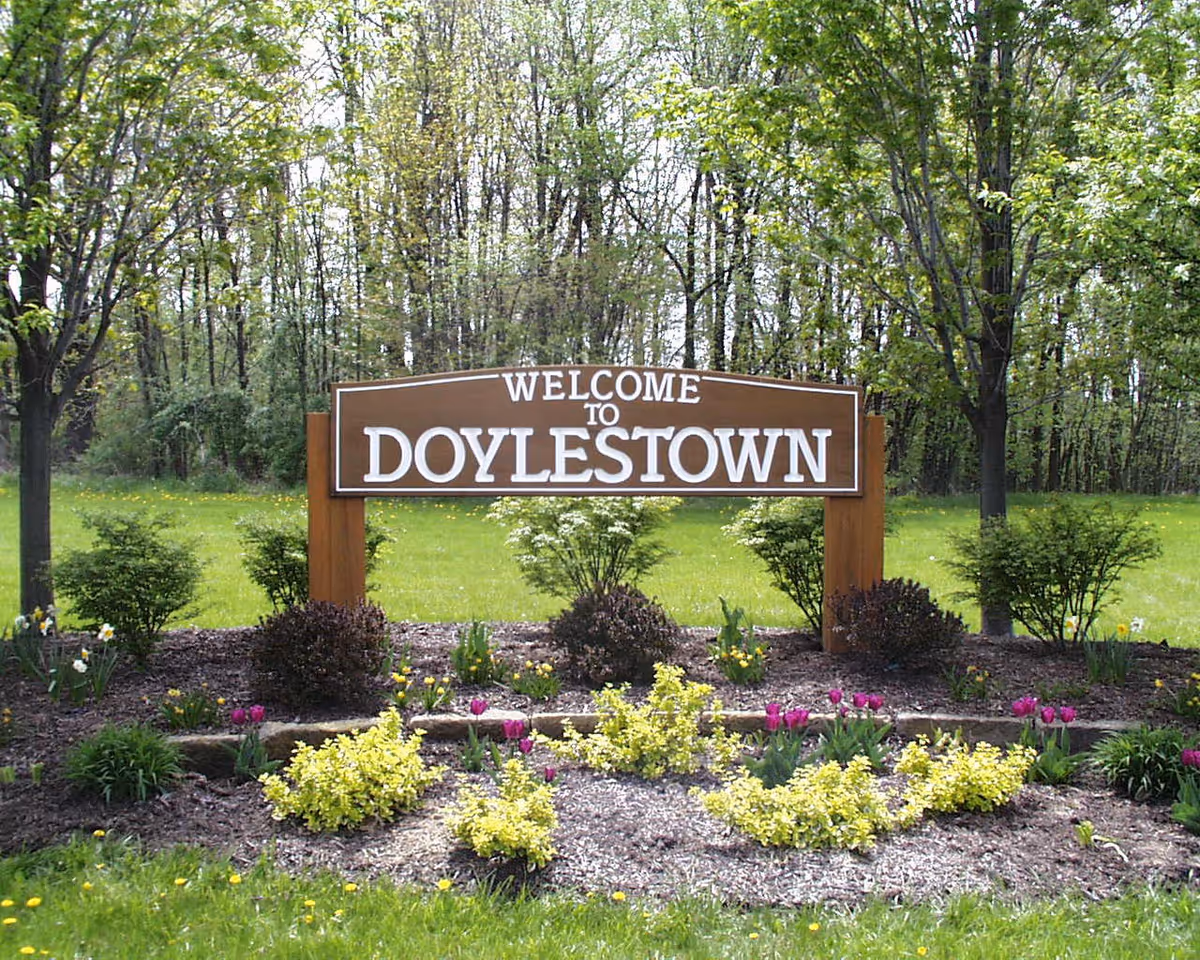A wooden sign that reads 'Welcome to Doylestown' surrounded by a landscaped garden with various bushes, flowers, and trees in the background.