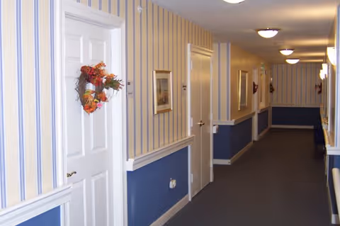 Carpeted interior hallway with striped wallpaper, white doors and wreaths, framed pictures and ceiling lights.