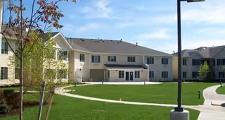 Exterior view of a senior living facility building with beige siding and multiple windows, surrounded by a well-maintained green lawn, young trees, and a paved walkway under a blue sky.