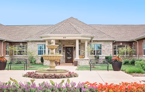Front exterior view of a single-story brick and stone building with a peaked roof, a central covered entrance with columns, a two-tiered stone fountain in front, benches on either side, and landscaped flower beds with colorful flowers and greenery.