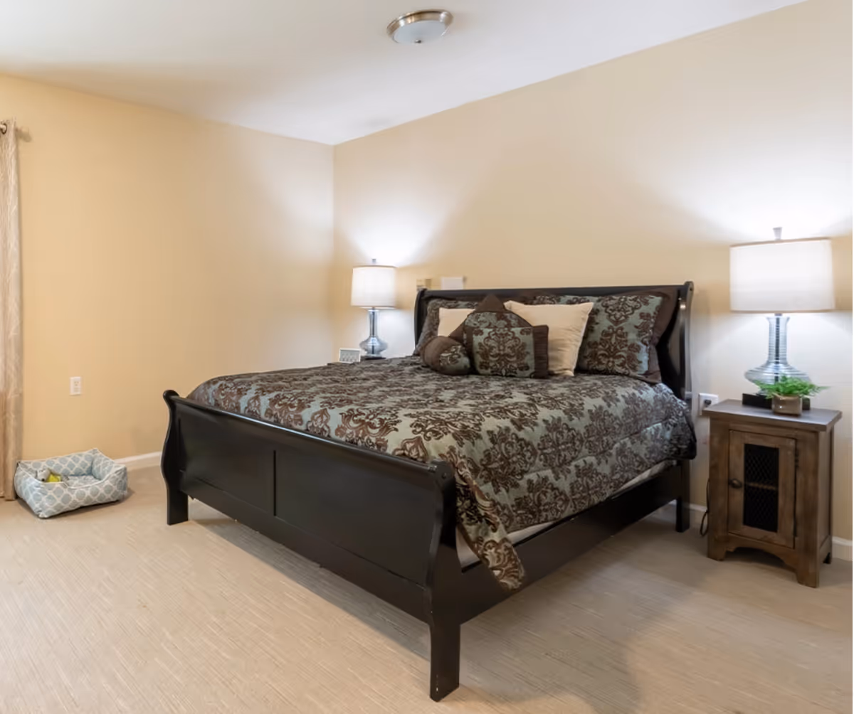 A bedroom with a dark wooden bed frame and a bed made with a patterned comforter and multiple pillows. There are two bedside tables, each with a lamp, and a small plant on the right table. A pet bed with a toy is placed on the carpeted floor near the left wall. The walls are painted beige and the room is softly lit.