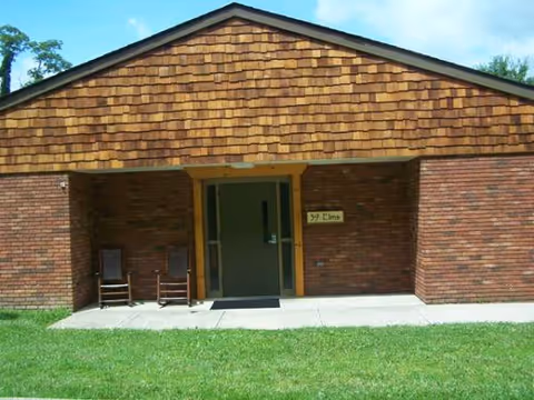 Brick building entrance with a shingled gable roof, two chairs on a small porch.