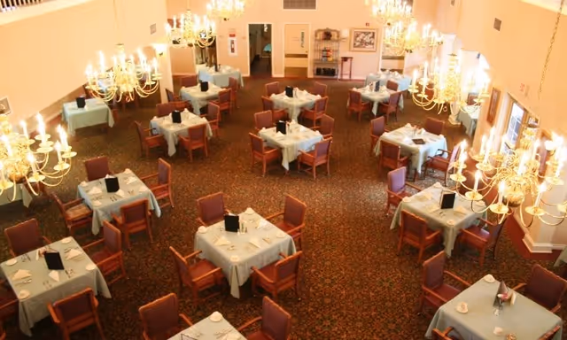 A spacious dining room with multiple square tables covered with light blue tablecloths, each surrounded by four wooden chairs with red cushions. The room is well-lit with several ornate chandeliers hanging from the ceiling. The carpet has a detailed pattern, and there are windows along one wall letting in natural light.