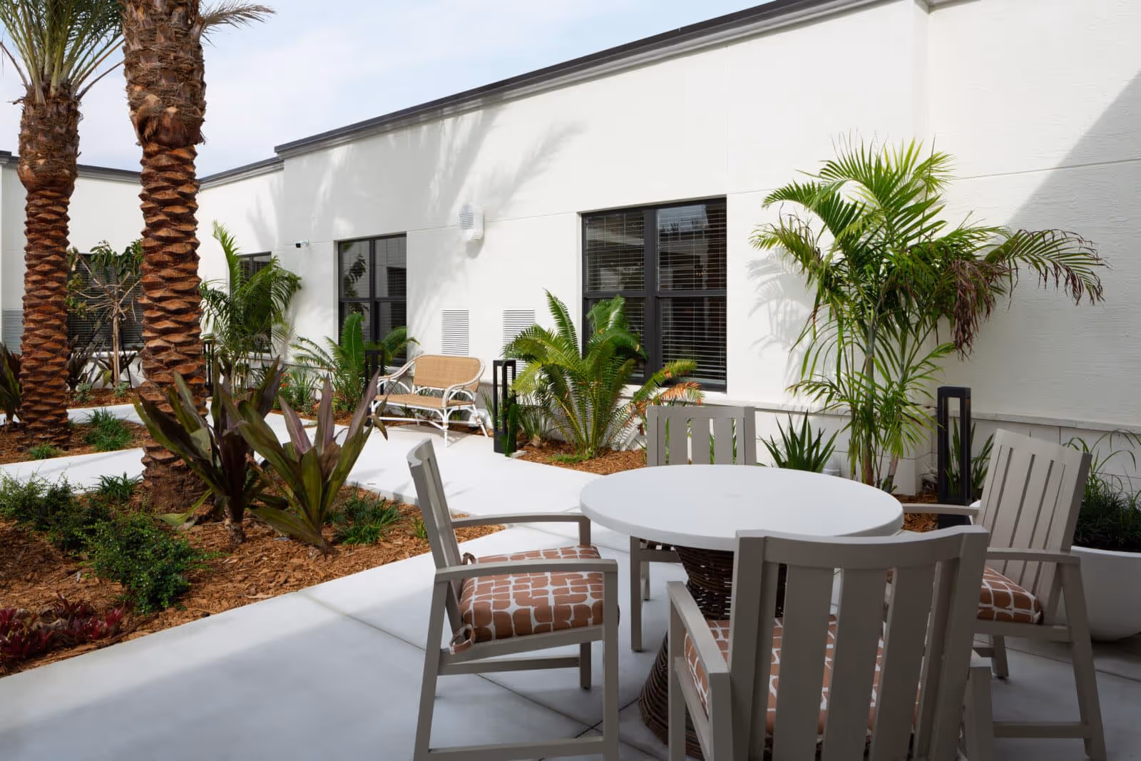 Outdoor patio area at The Palms at Plantation featuring a round white table with four cushioned chairs, surrounded by various tropical plants and palm trees, with a white building wall and windows in the background.
