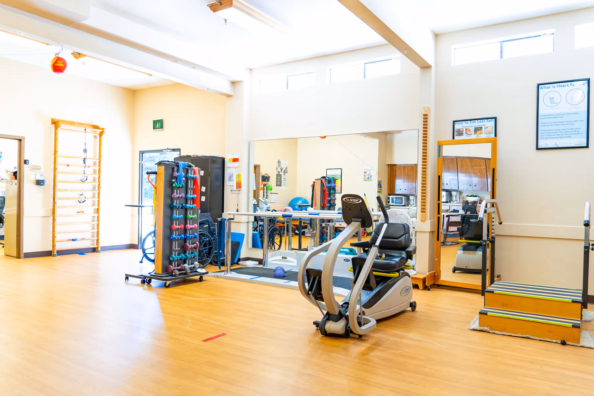 Bright rehabilitation/exercise room with fitness equipment, hand weights, mirrors and wooden flooring.