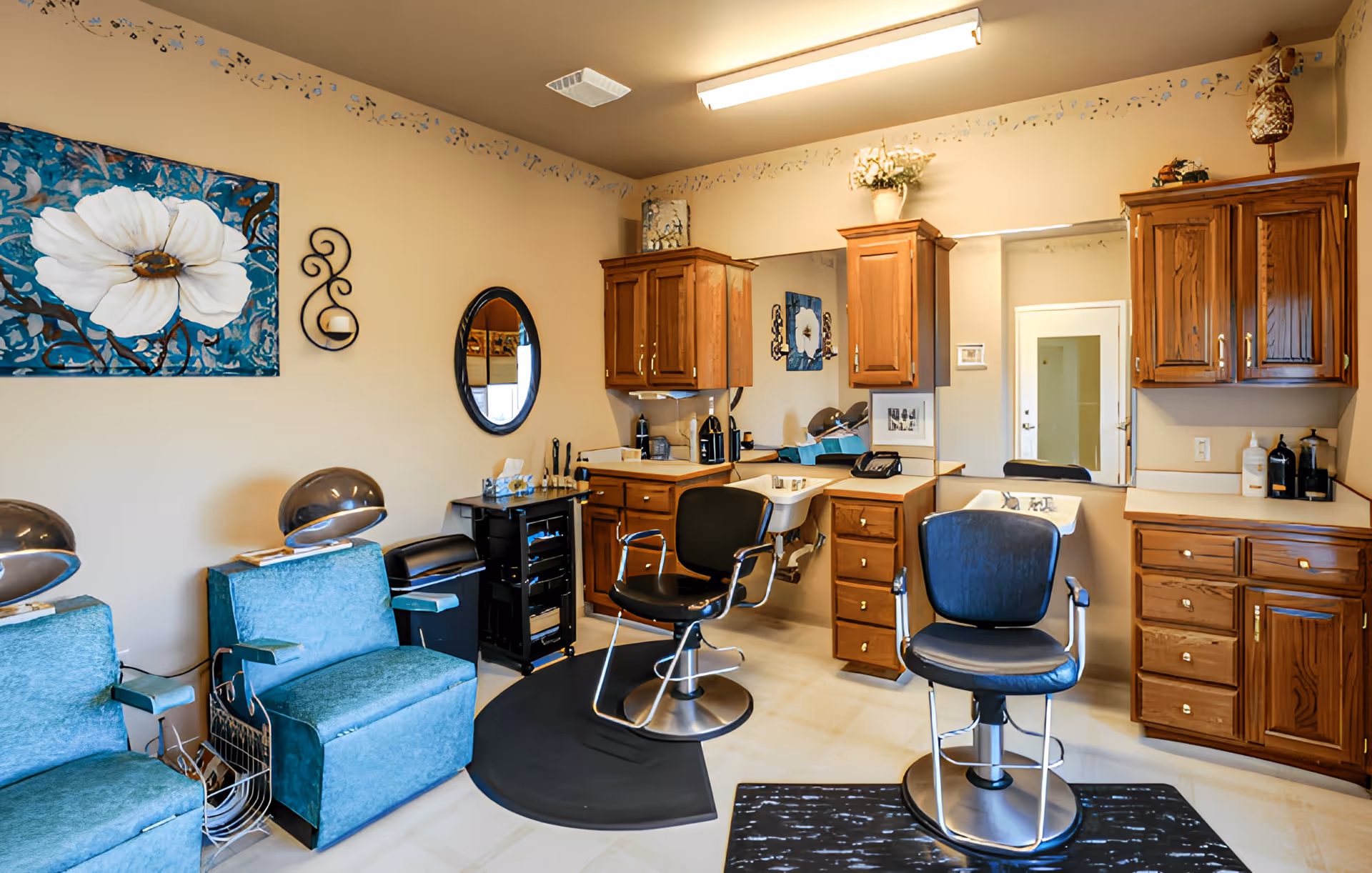 Interior of a hair salon area in a senior living facility with two black salon chairs in front of sinks and wooden cabinets. There are two blue upholstered chairs with hair dryers on the left side, a large mirror on the wall, and a painting of a white flower on a blue background.