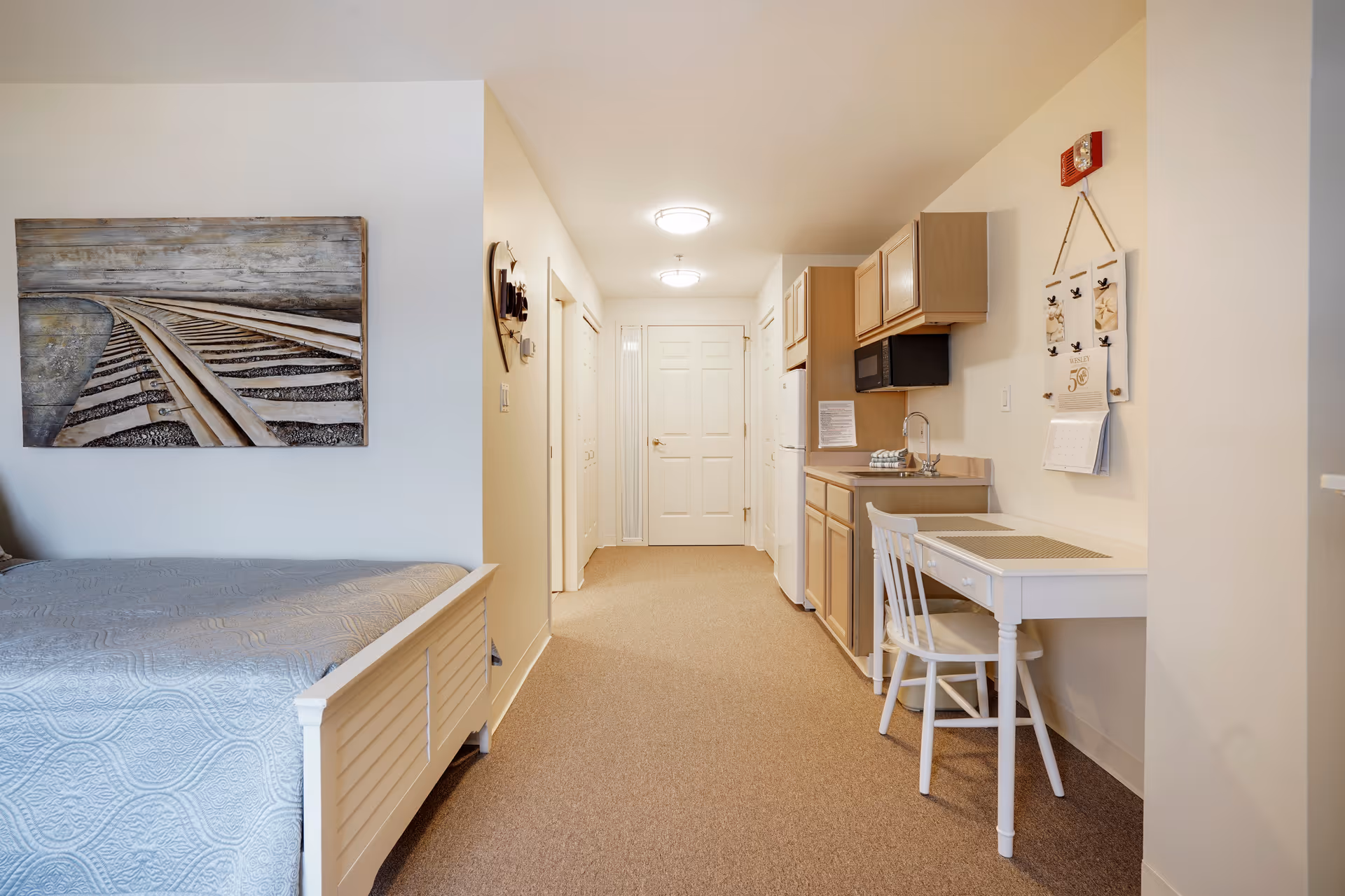 Interior view of a senior living facility room at Wesley Ridge featuring a bed with a light blue quilt, a painting of railroad tracks on the wall, a small kitchenette with wooden cabinets, a white refrigerator, microwave, sink, and a small white table with a chair. The hallway leads to a closed door at the end.