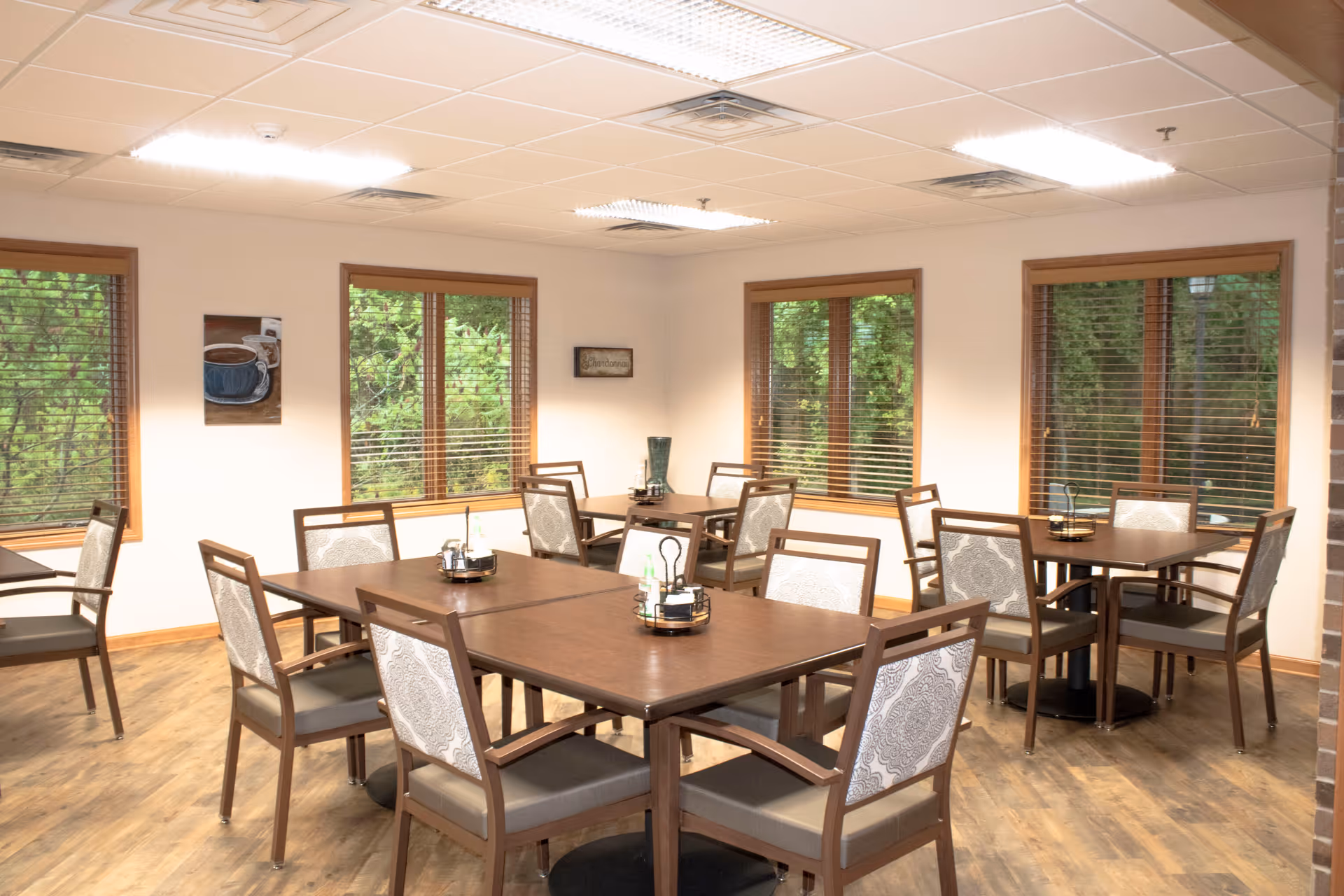 A bright dining room with multiple wooden tables and cushioned chairs arranged neatly. The room has large windows with wooden blinds, allowing natural light to illuminate the space. The walls are light-colored, and there are a few decorative items including a painting of a coffee cup and a small wall sign. The floor is wooden, and the ceiling has recessed lighting.