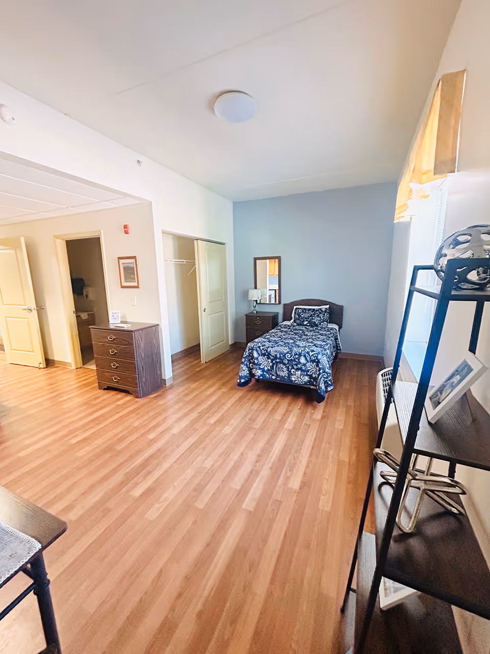 A spacious bedroom with wooden flooring, a single bed with a blue floral bedspread, a nightstand with a lamp and mirror, an open closet with sliding doors, a chest of drawers, and a shelving unit near a window with a yellow curtain.