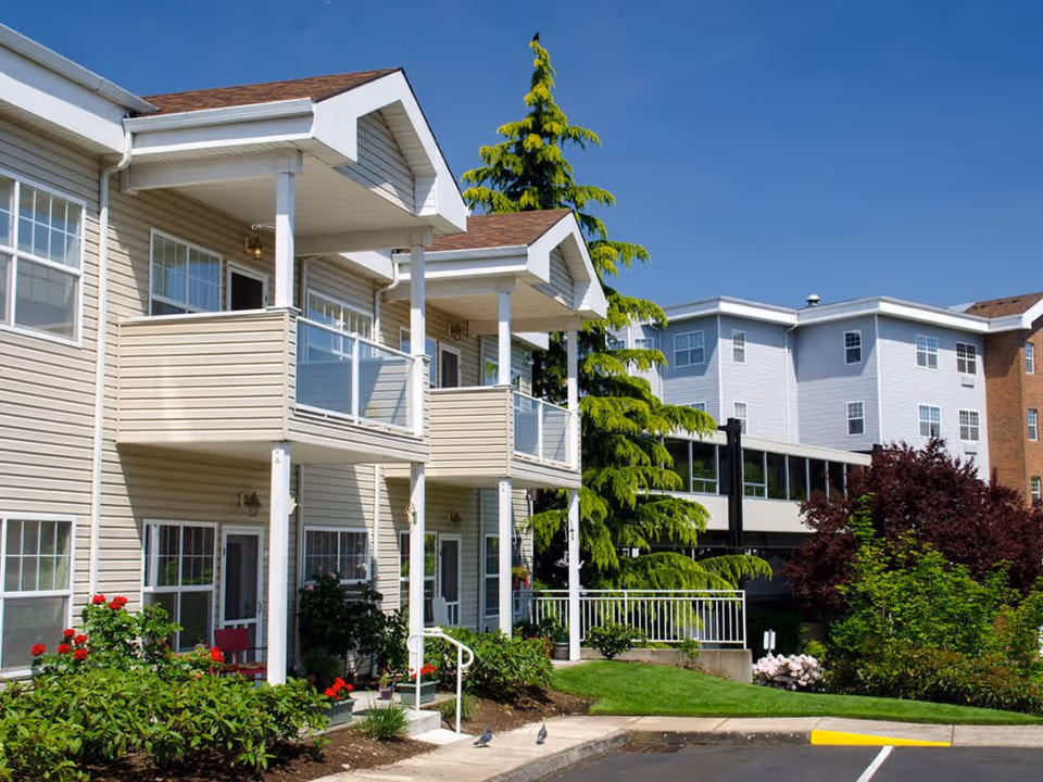 Exterior view of a senior living facility with beige siding and multiple balconies. The building is surrounded by well-maintained landscaping including bushes, flowers, and trees under a clear blue sky.