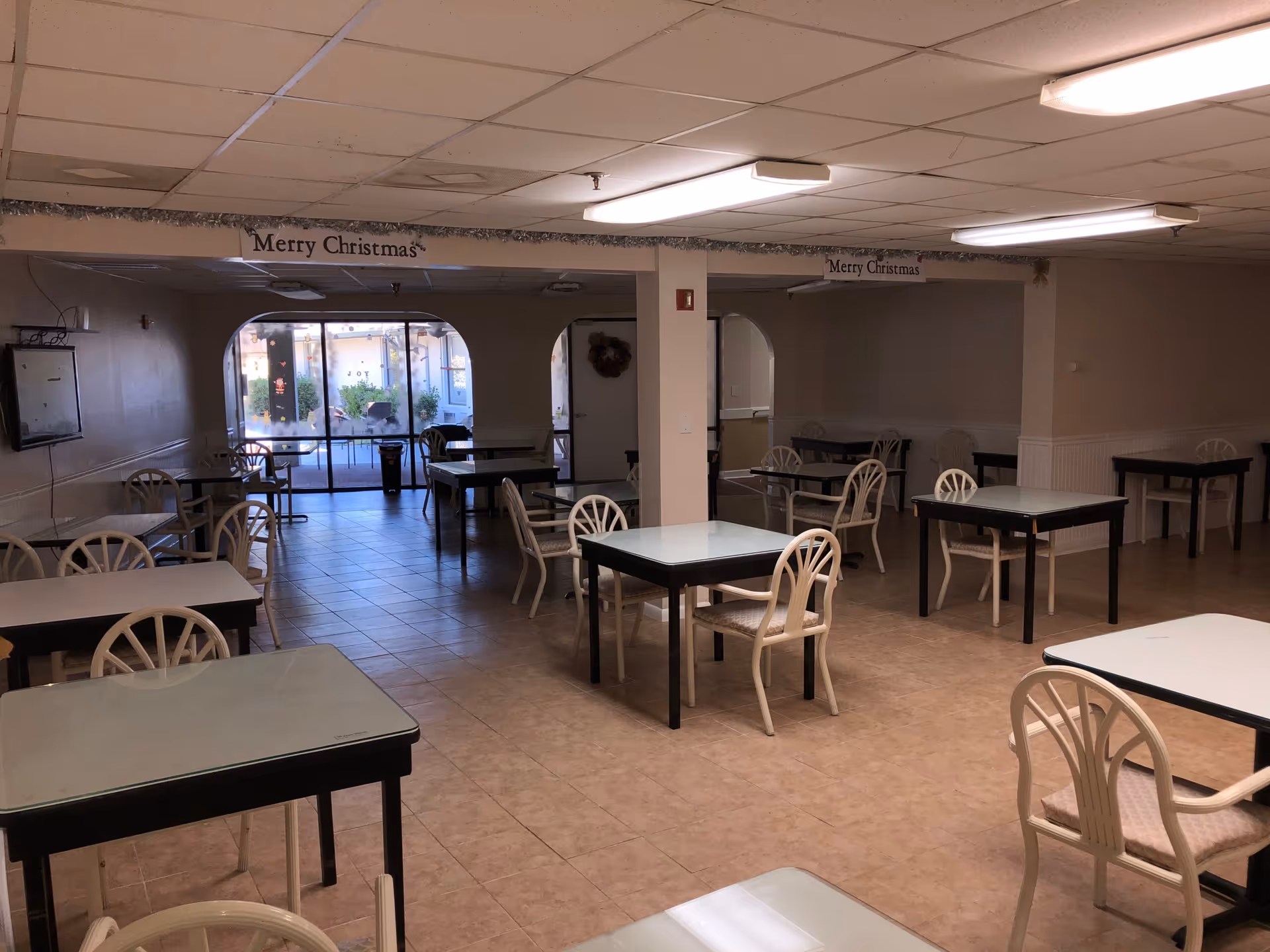 Interior view of a dining room with multiple square tables and chairs arranged neatly. The room has tiled flooring, a drop ceiling with fluorescent lights, and a glass door leading to an outdoor area. There are 'Merry Christmas' signs hanging from the ceiling beams.