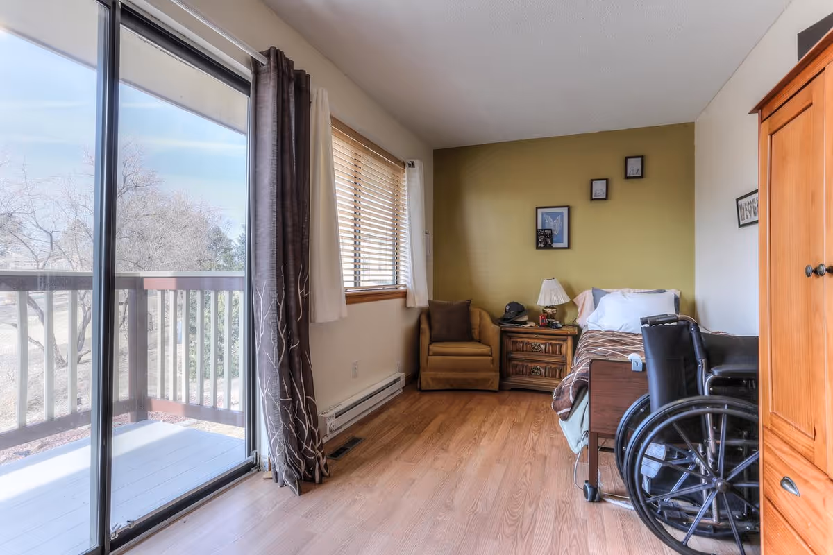 A bright senior bedroom with a single bed, wheelchair, armchair, nightstand, and sliding glass doors opening to a balcony.