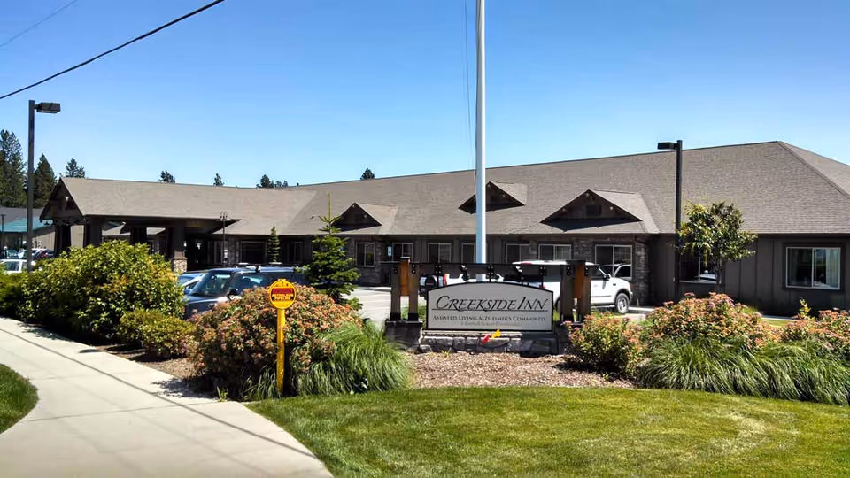Exterior view of Creekside Inn Memory Care Community building with a clear blue sky, landscaped bushes, a sidewalk, parked cars, and a sign in front that reads 'Creekside Inn Assisted Living Alzheimer's Community'.