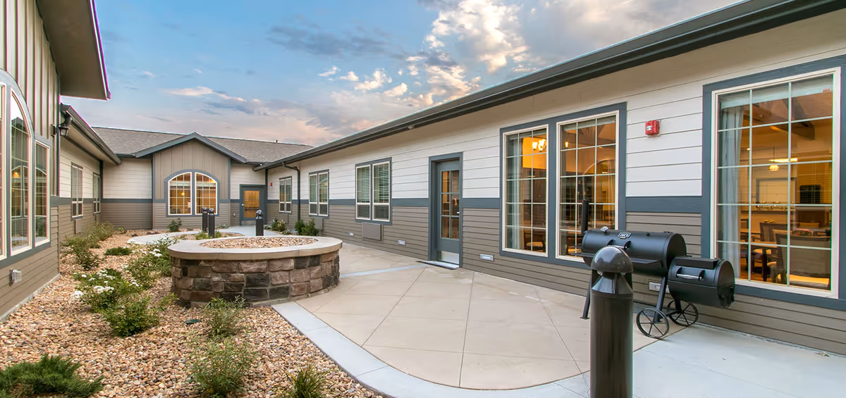 Courtyard with a circular stone firepit, gravel landscaping, a smoker grill, and exterior windows and doors of a single-story senior living building.