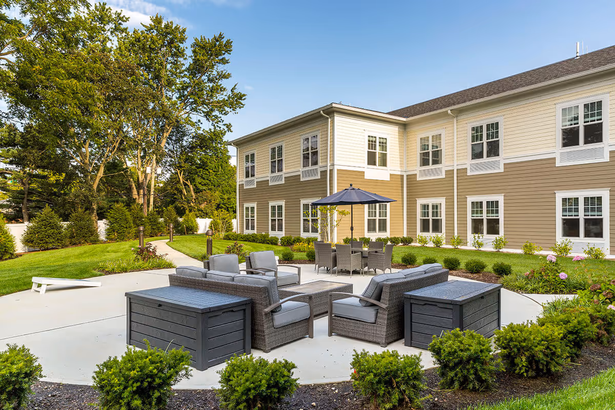 Outdoor patio area at Orchard Estate of Woodbury featuring comfortable wicker seating with cushions, a table with an umbrella, surrounded by well-maintained landscaping and greenery, adjacent to a two-story beige and white building under a clear blue sky.