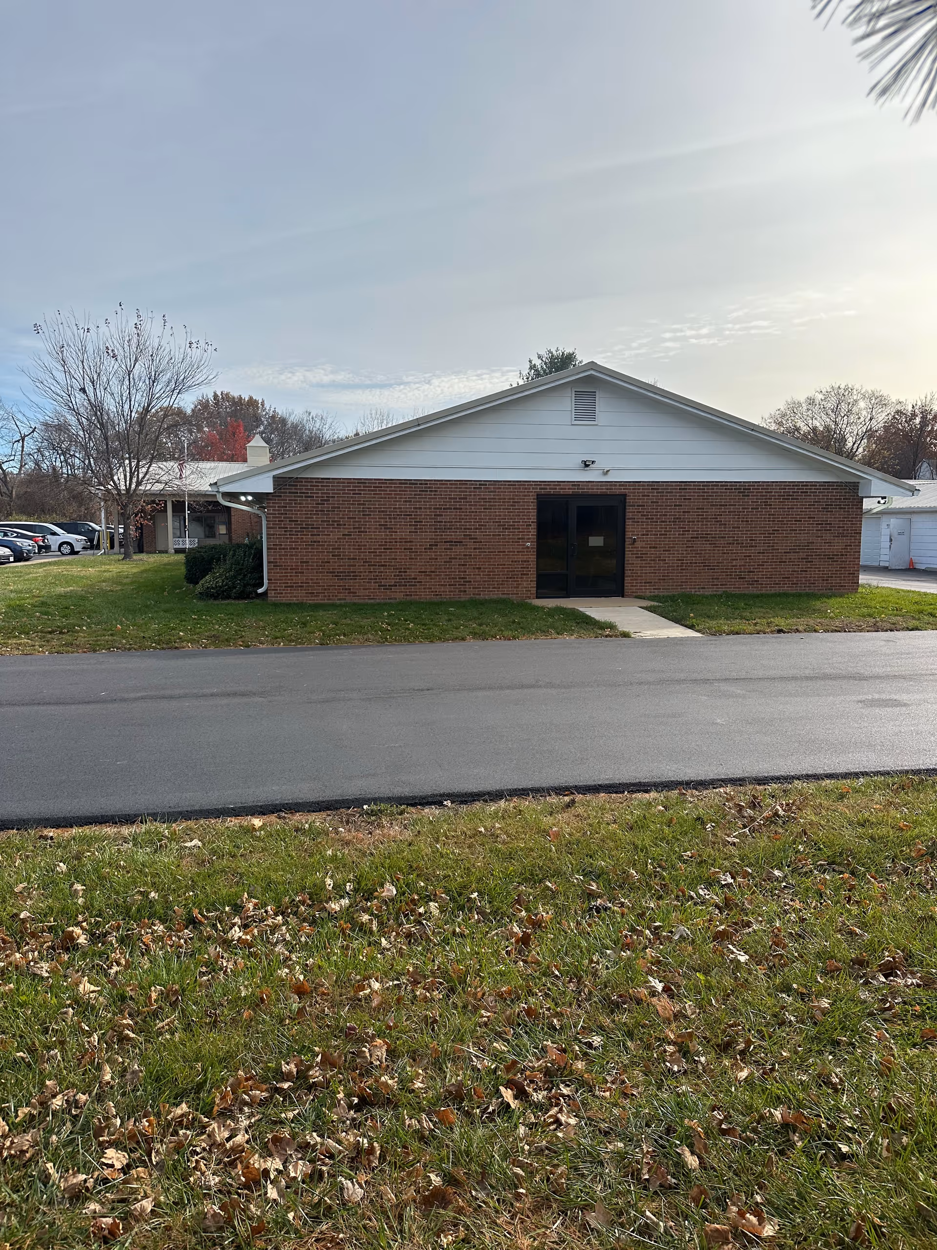 Exterior view of a single-story brick building with a white gabled roof and a glass door entrance. The building is surrounded by grass with some fallen leaves and a paved road in front. Trees with autumn foliage are visible in the background under a partly cloudy sky.