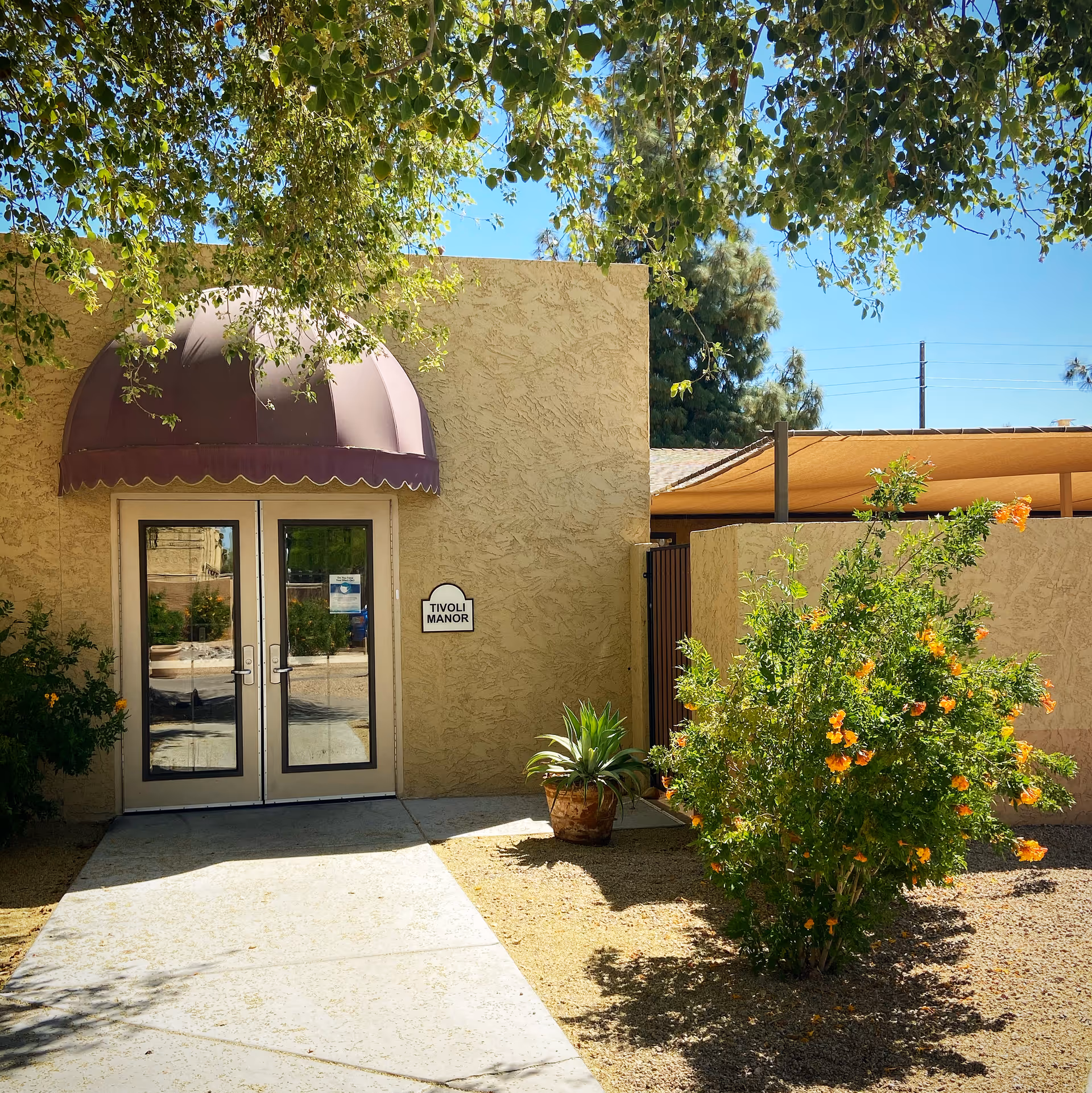 Exterior view of a building entrance with double glass doors under a brown awning. A sign next to the door reads 'Tivoli Manor'. There are green plants and a flowering bush with orange flowers in front of the building, and a tree with green leaves partially shading the area.