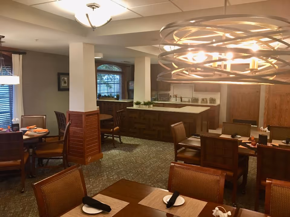 Interior dining room with multiple wooden tables and chairs, a large decorative chandelier, and a serving counter in the background.