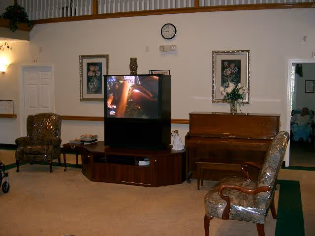 Interior view of a living room area in a senior living facility with a television on a wooden stand, two upholstered chairs, a piano with a vase of flowers on top, framed floral artwork on the walls, a clock above the TV, and a doorway leading to another room.
