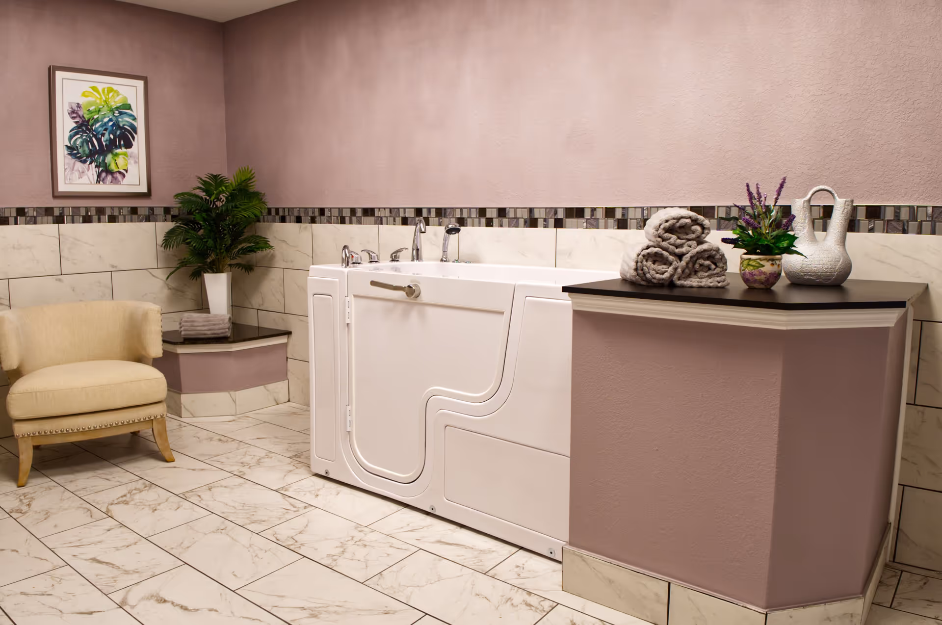 A spacious bathroom with a walk-in bathtub featuring a door and multiple faucets. The walls are tiled halfway up with marble tiles and a decorative mosaic border, while the upper walls are painted a soft mauve color. There is a beige upholstered chair with wooden legs on the left, a potted green plant in the corner, and a framed botanical artwork on the wall. On the right, a countertop holds three rolled gray towels, a small potted plant with purple flowers, and a white decorative vase.
