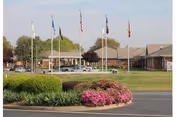 Front view of a single-story brick senior living facility with multiple flagpoles and landscaped shrubs and flowering beds.
