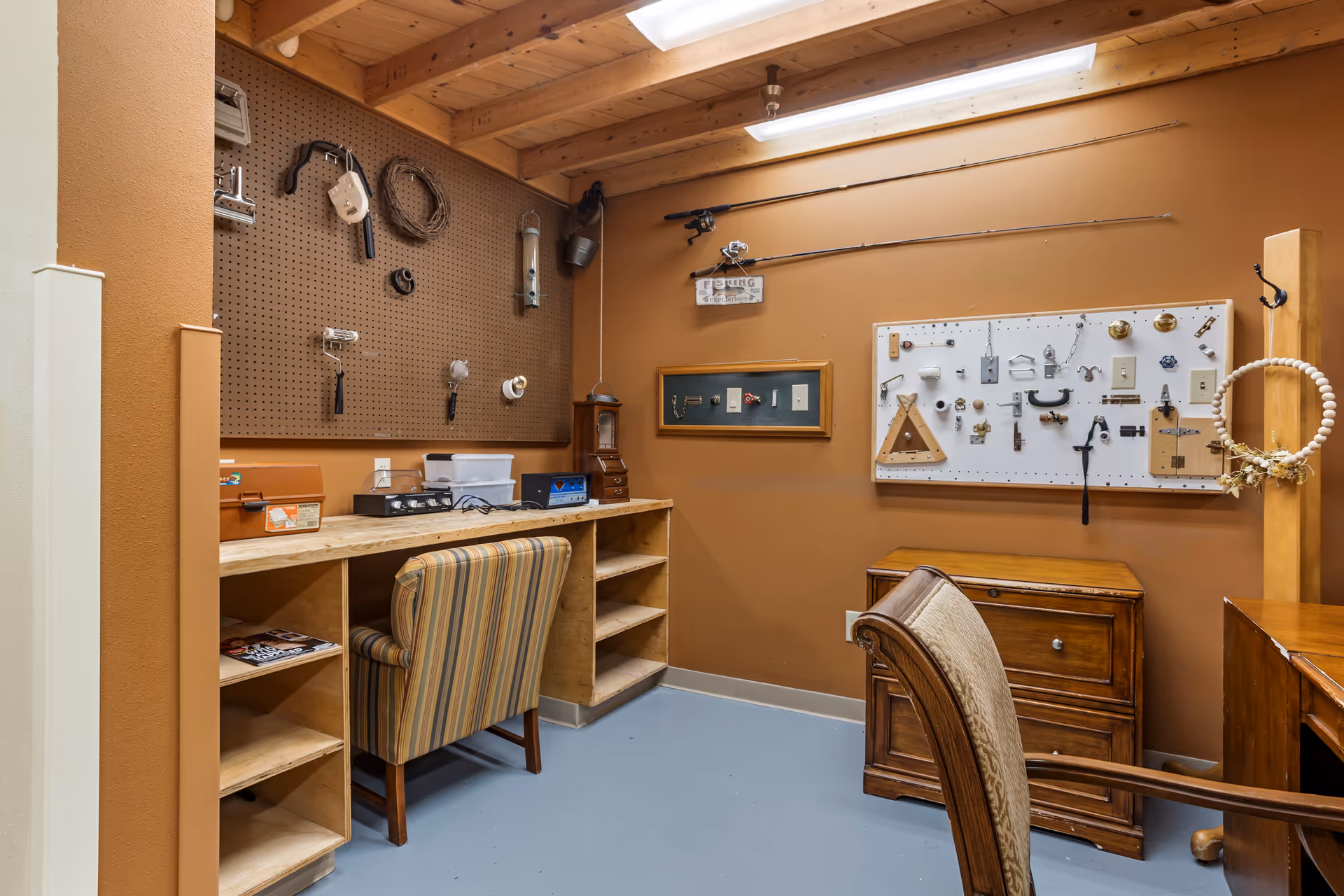 A small workshop or craft room with brown walls and a wooden ceiling. There is a long wooden desk with a striped upholstered chair in front of it. On the wall above the desk is a pegboard with various tools and items hanging. Another board on the adjacent wall displays different hardware pieces like locks, handles, and switches. Two fishing rods are mounted on the wall above a small wooden cabinet. The floor is painted light blue.