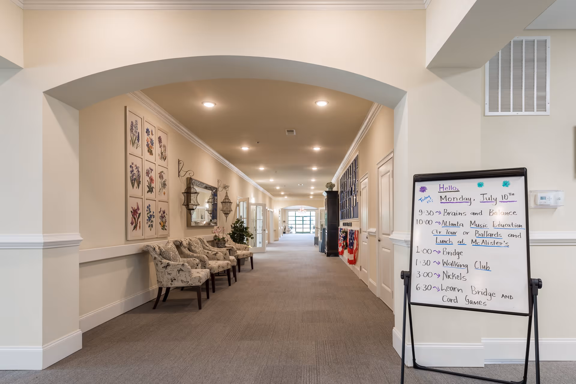 A well-lit hallway in a senior living facility with beige walls and carpeted floor. On the left side, there are four upholstered chairs with floral patterns beneath framed botanical artwork and decorative wall sconces. On the right side, a whiteboard displays the day's schedule of activities including music education, bridge, walking club, and card games. The hallway extends into the distance with doors and windows along the sides.