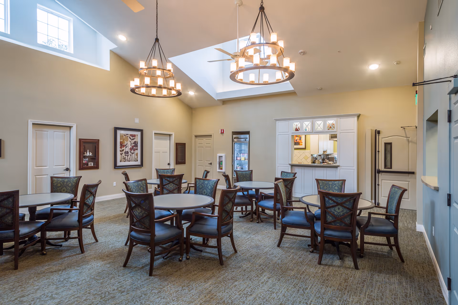 A well-lit dining area in a senior living facility with multiple round tables surrounded by wooden chairs with patterned cushions. The room features high ceilings with two large circular chandeliers, beige walls, and carpeted floors. There is a pass-through window to a kitchen area and several closed doors along the walls.