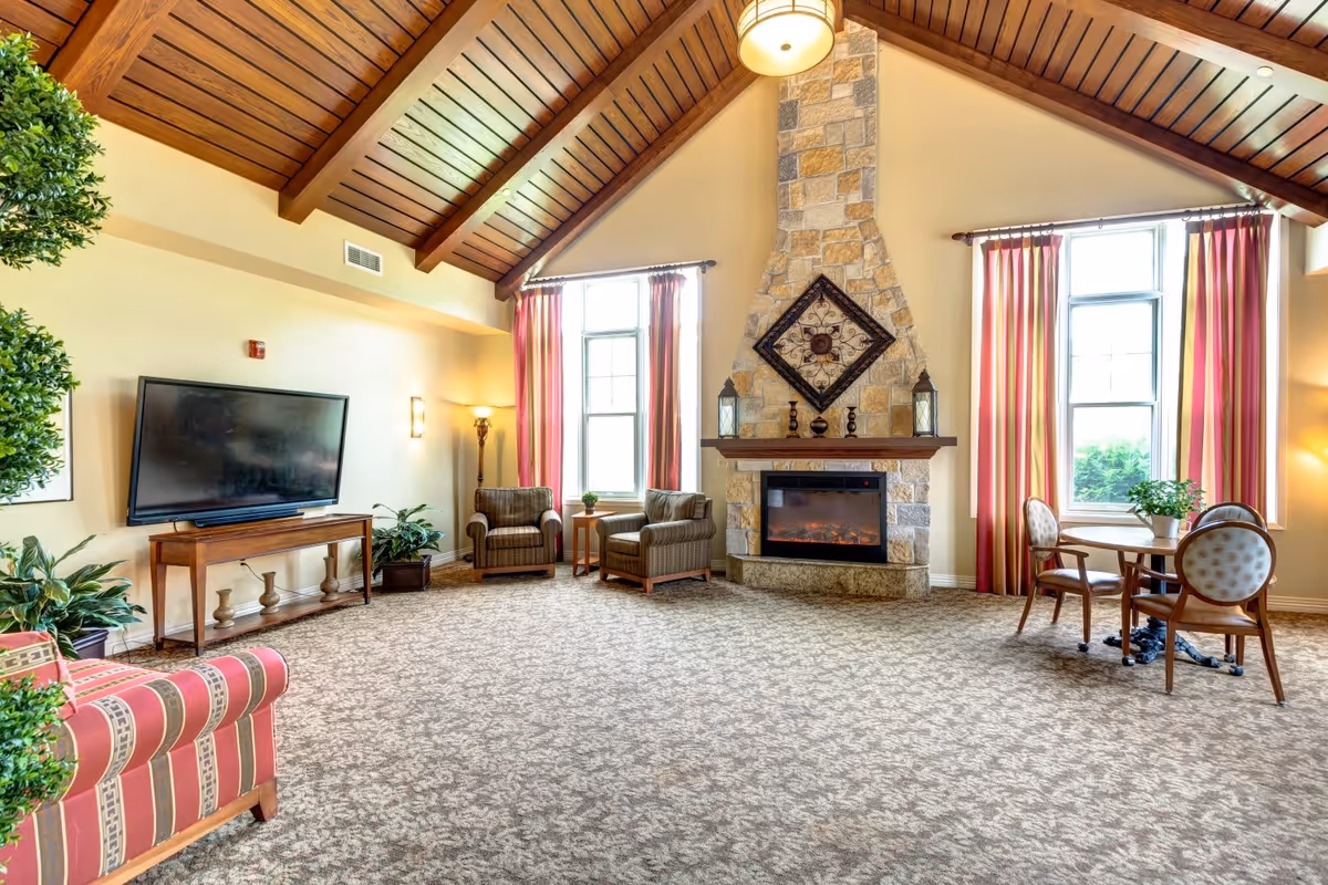 A spacious living room with a high wooden vaulted ceiling, a stone fireplace with decorative items on the mantel, two large windows with red and beige striped curtains, two armchairs with a small table between them, a round table with three chairs, a flat-screen TV on a wooden stand, and several potted plants.