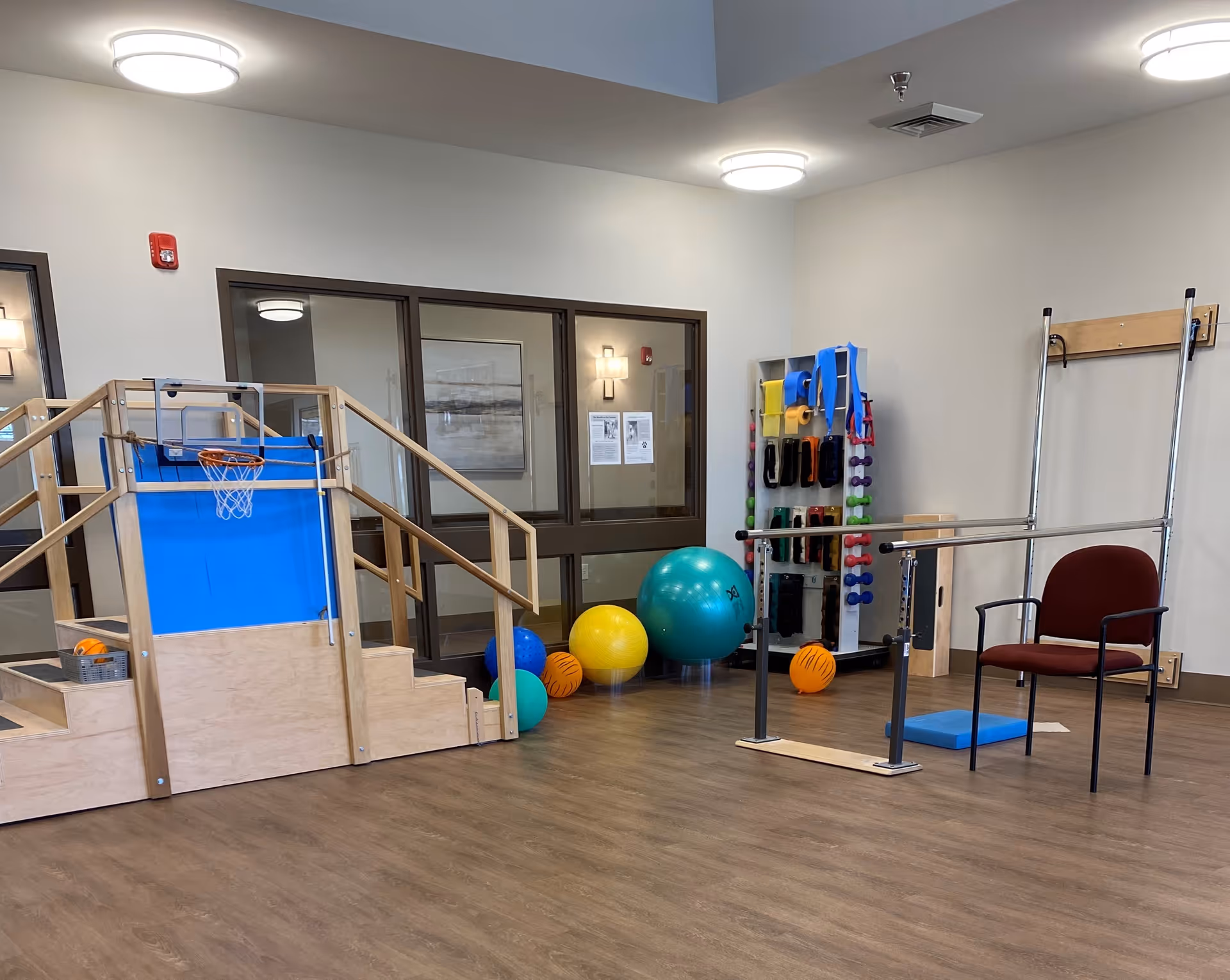 A physical therapy or exercise room in an assisted living facility with a small wooden staircase and ramp with a basketball hoop, various exercise balls, colorful dumbbells on a rack, a chair, and parallel bars for walking support.