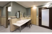 A clean and modern bathroom area with a long white sink countertop, a round mirror mounted on the wall, a chair placed in front of the sink, and a wooden door with frosted glass. The walls are painted in muted green and beige tones with decorative tile accents.