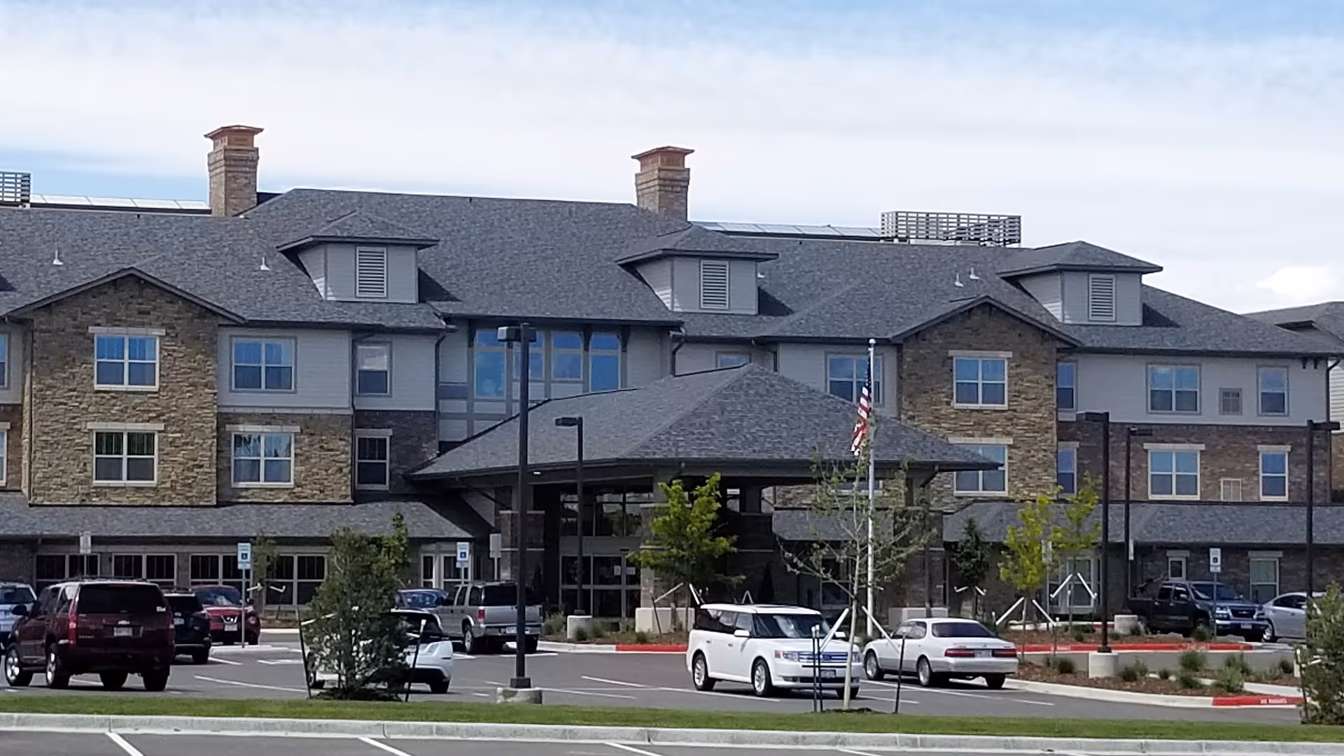 Front exterior of a multi-story senior living building with a covered entrance, parked cars, and a landscaped parking lot.