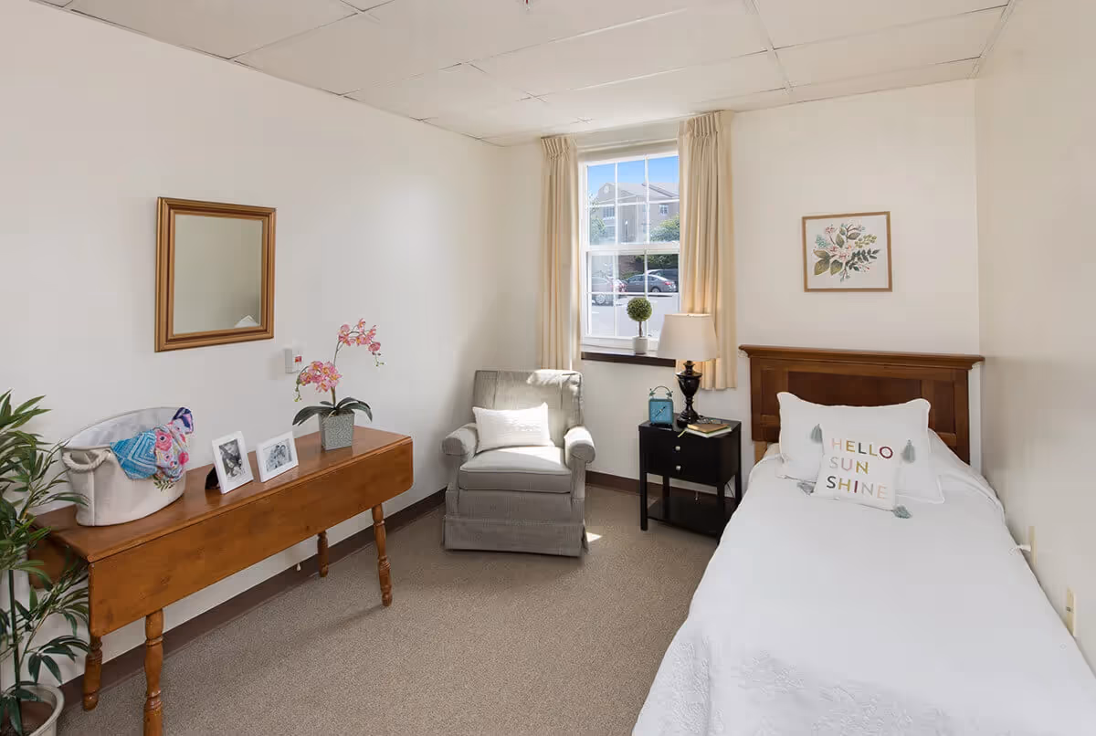 A small, cozy bedroom with a single wooden bed featuring a white bedspread and a decorative pillow that says 'HELLO SUNSHINE'. Next to the bed is a black nightstand with a lamp, a small blue lantern, and a book. A gray armchair with a white pillow is positioned near a window with cream-colored curtains, letting in natural light. On the opposite wall, there is a wooden table with a potted orchid, two framed photos, and a basket with colorful textiles. A green potted plant is on the floor beside the table. The walls are white, and there is a framed floral artwork above the bed.
