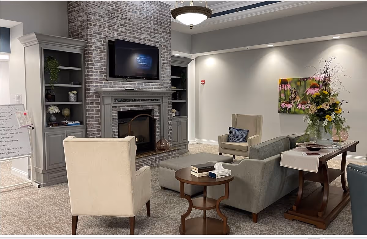 A cozy living room area with a gray brick fireplace and a mounted flat-screen TV above it. The room features two beige armchairs, a gray sofa, a round wooden side table with books and tissues, and a wooden console table with a vase of flowers. Built-in gray shelves flank the fireplace, and a colorful floral painting hangs on the wall. The space is softly lit with ceiling lights and a hanging light fixture.