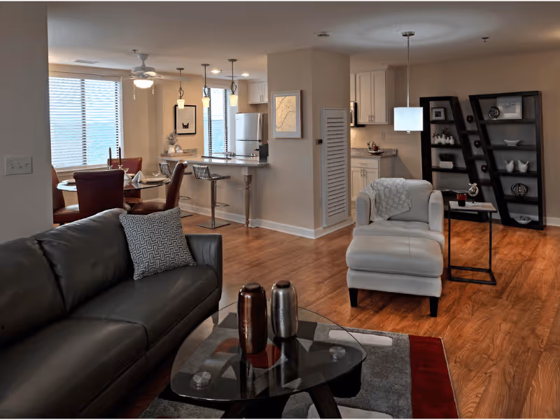 A modern living area featuring a dark leather sofa with a patterned pillow, a glass coffee table with decorative vases, a white armchair with a throw blanket, and a side table with a drink. In the background, there is a kitchen with white cabinets, a refrigerator, and a breakfast bar with two stools. To the left, a dining area with a round table and red chairs is visible near large windows with blinds. The room has wooden flooring and neutral-colored walls with framed artwork and shelves displaying decorative items.