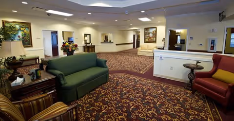 Spacious senior living facility common area with patterned carpet, multiple seating options including green, red, and beige armchairs and sofas, side tables with lamps and flowers, and framed artwork on the walls under bright ceiling lights.